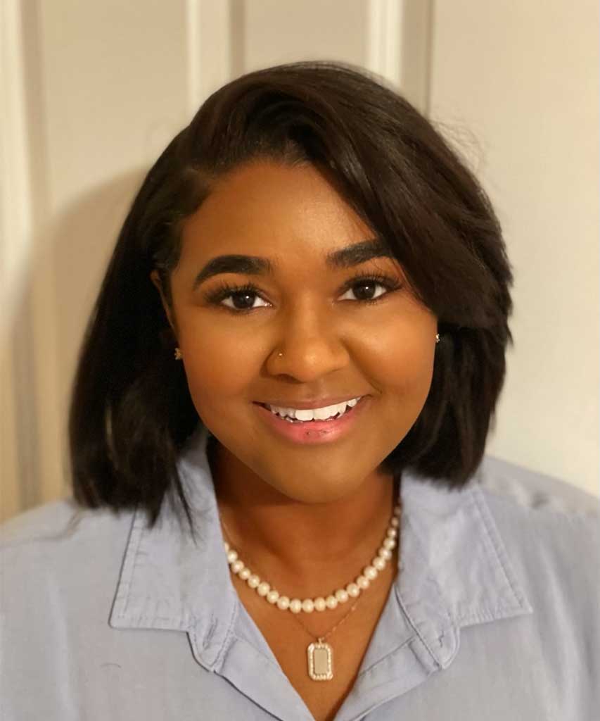 Smiling woman with straight, shoulder-length dark hair, wearing a light blue collared shirt, a pearl necklace, and a pendant, standing in front of a neutral background.