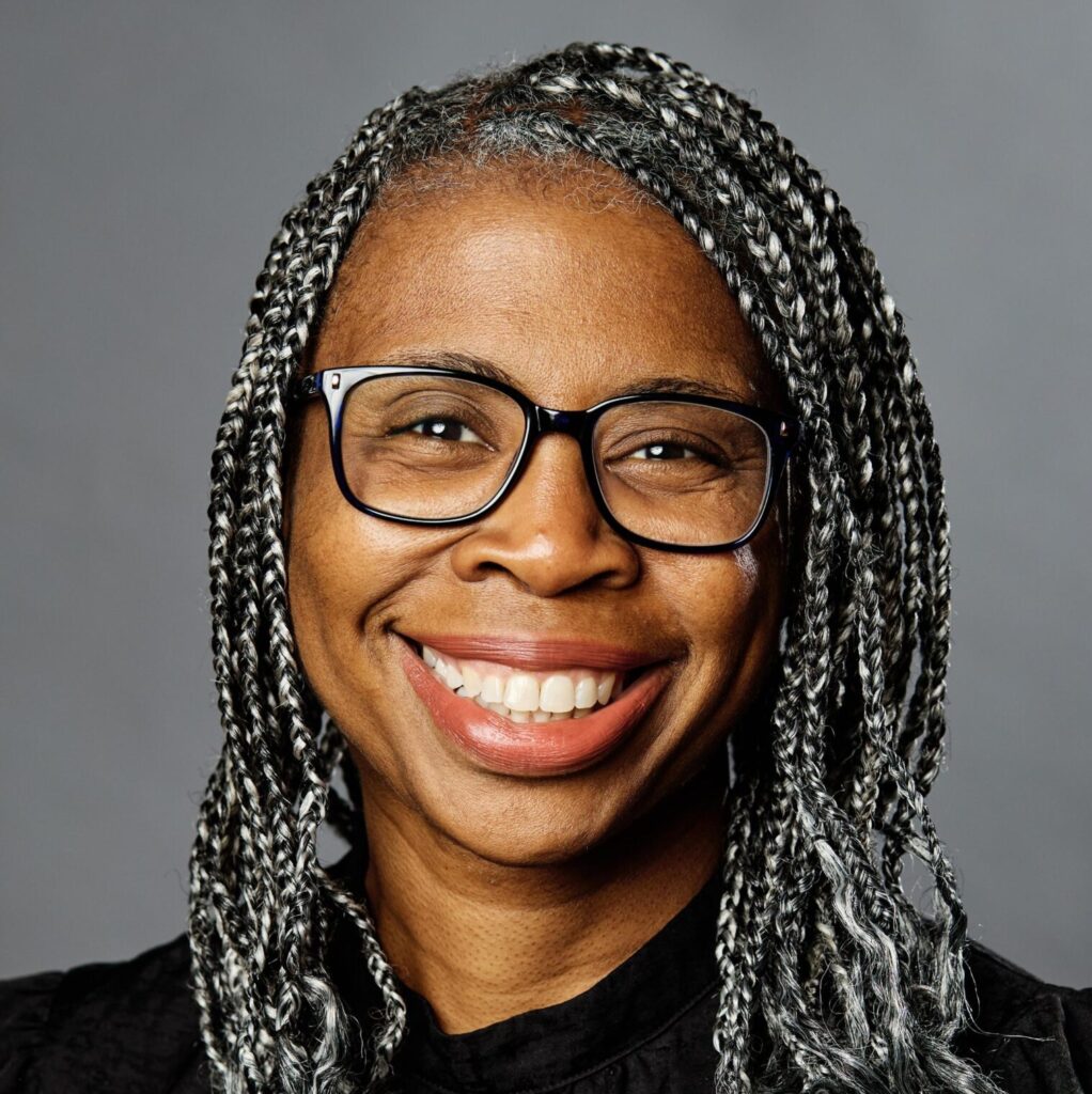 A woman with glasses and long, gray-and-black braided hair smiles at the camera. She is wearing a black top and is posed in front of a plain gray background.