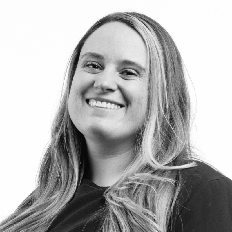 Black and white portrait of a woman with long, light hair wearing a dark top, smiling and looking slightly off to the side against a plain white background.
