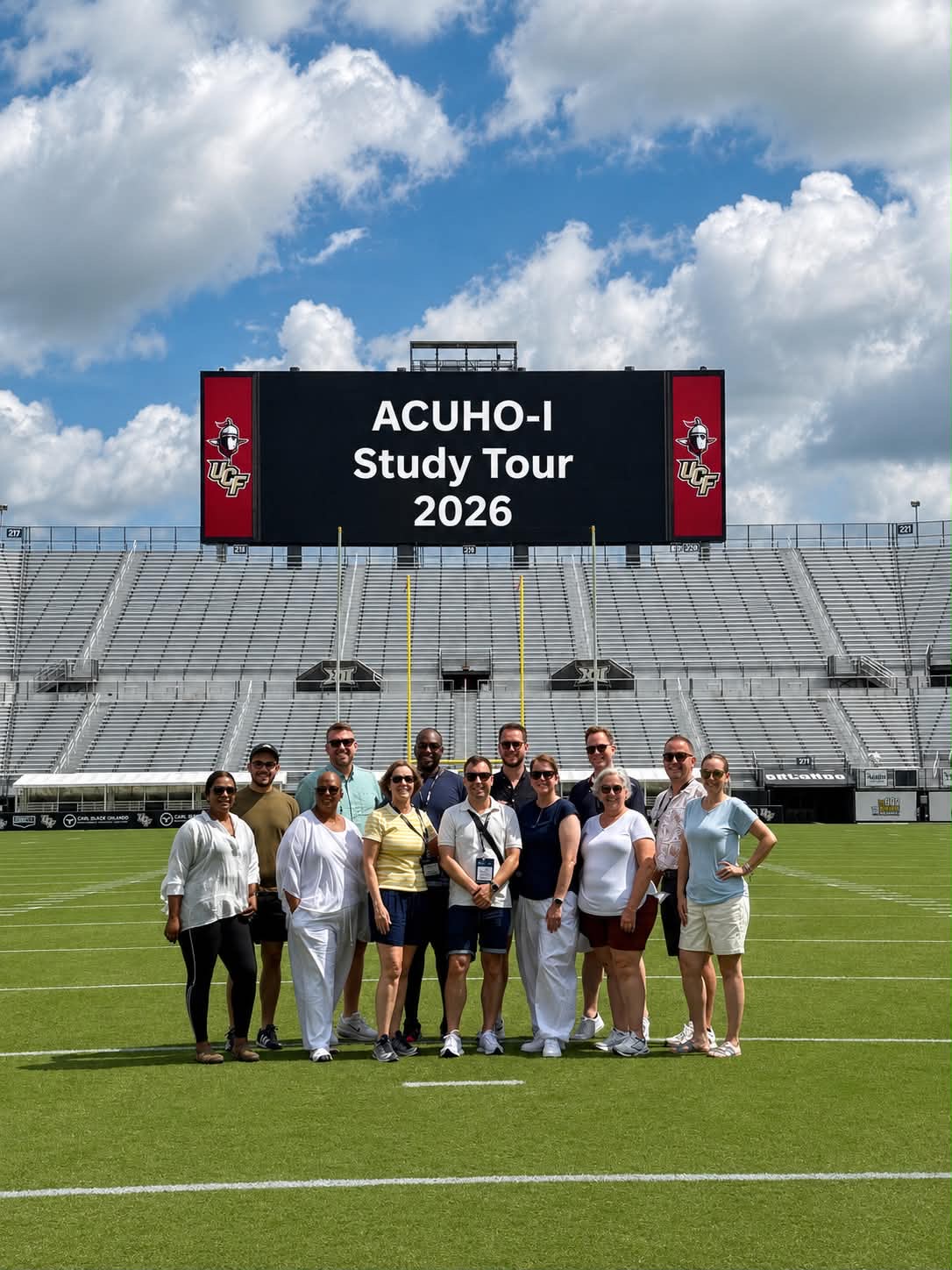 A group of people standing on a football field in front of a stadium scoreboard that reads ACUHO-I Study Tour 2026.