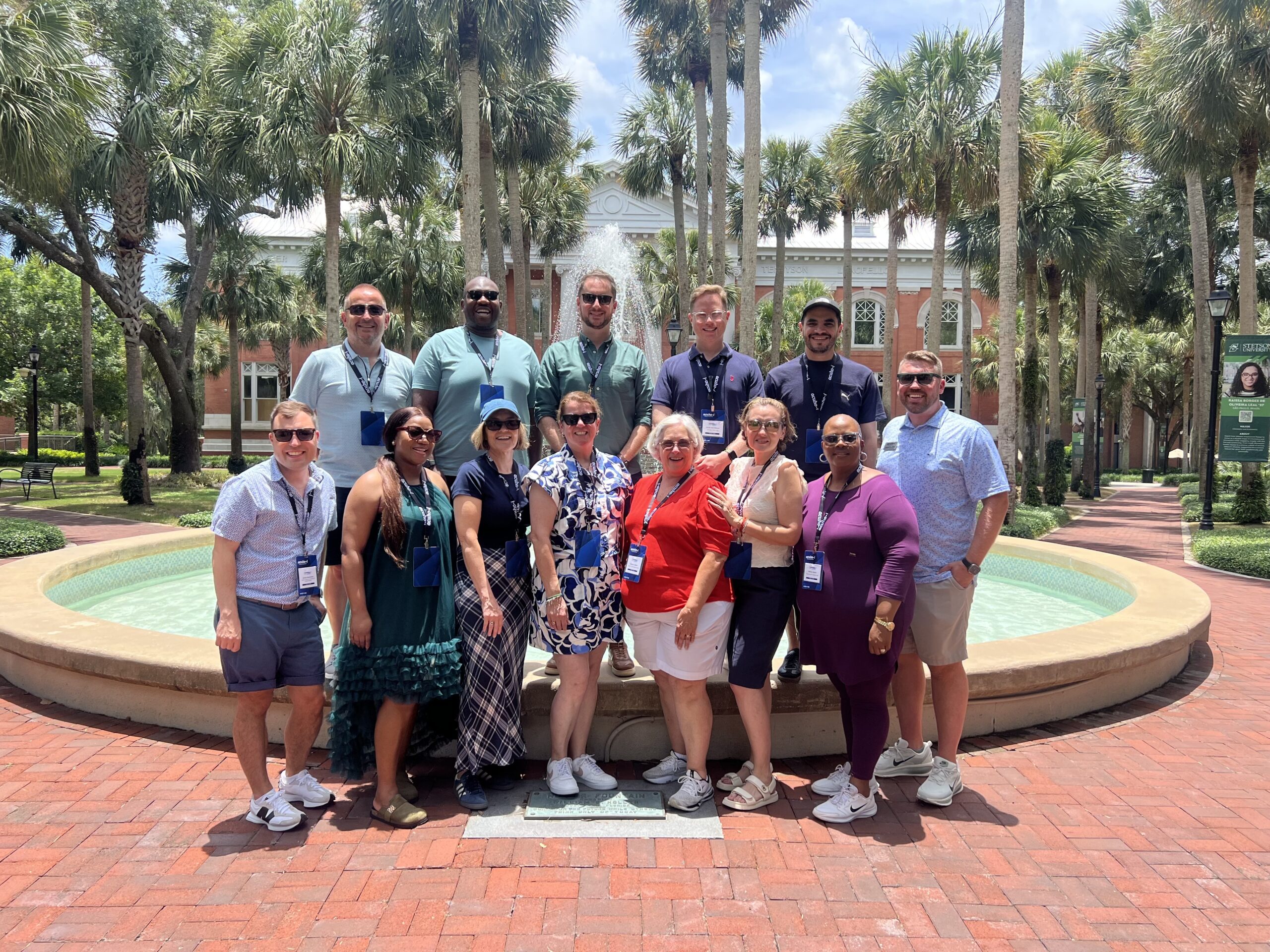 A group of fourteen adults wearing conference lanyards pose for a photo in front of a fountain and palm trees.