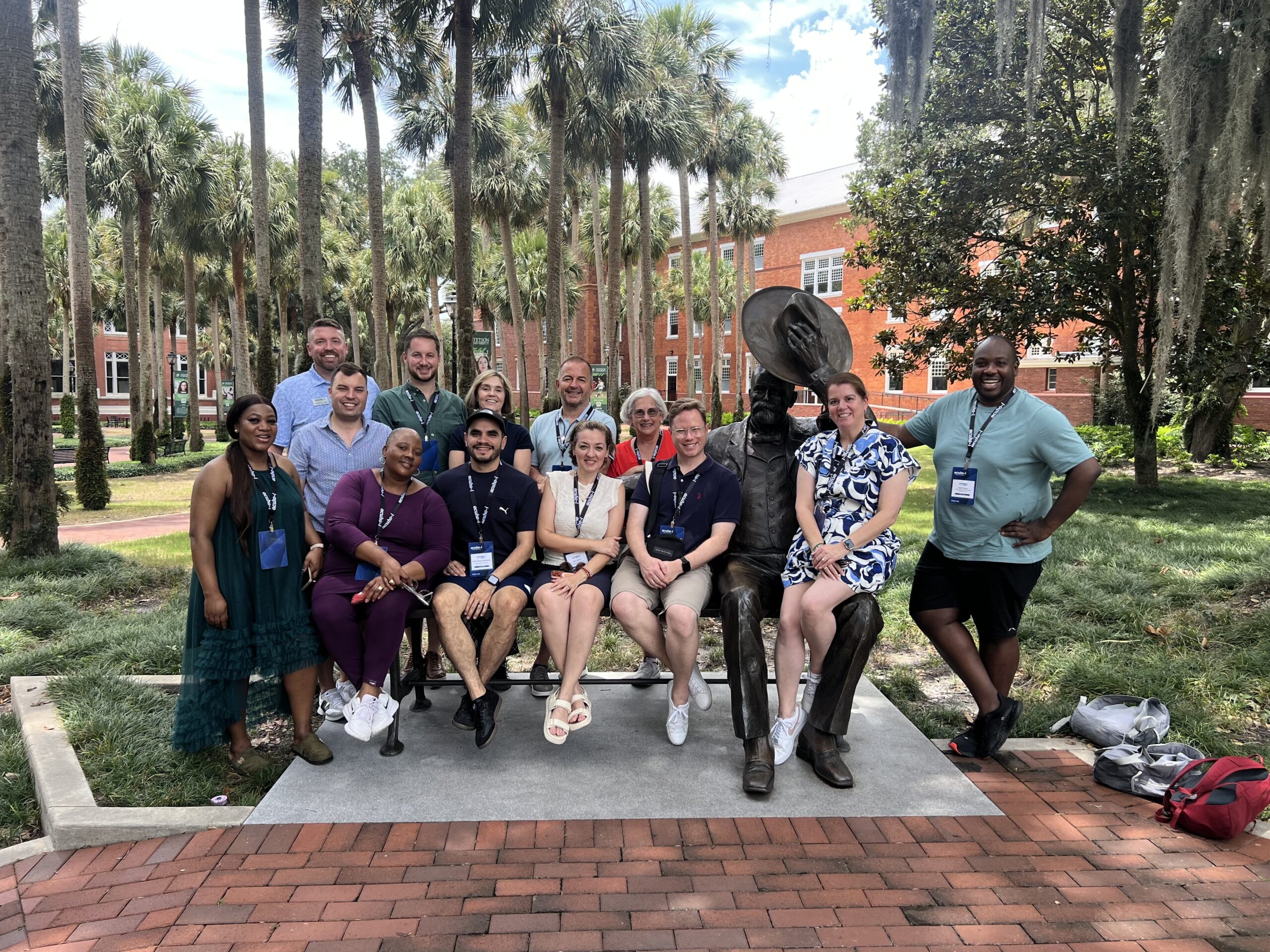 A group of thirteen people posing for a photo with a bronze statue of a man holding a hat on a bench in a park.