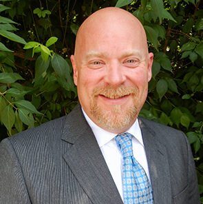 A smiling bald man with a goatee wearing a grey suit, white shirt, and blue patterned tie, posed in front of green foliage.